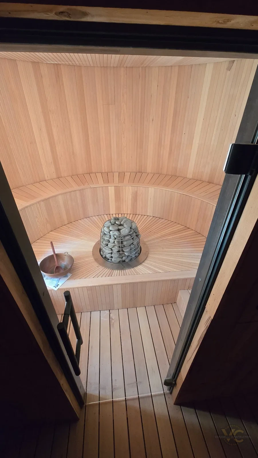 Finished Applegate Valley round sauna viewed through the glass door with curved wood seating and central stone heater